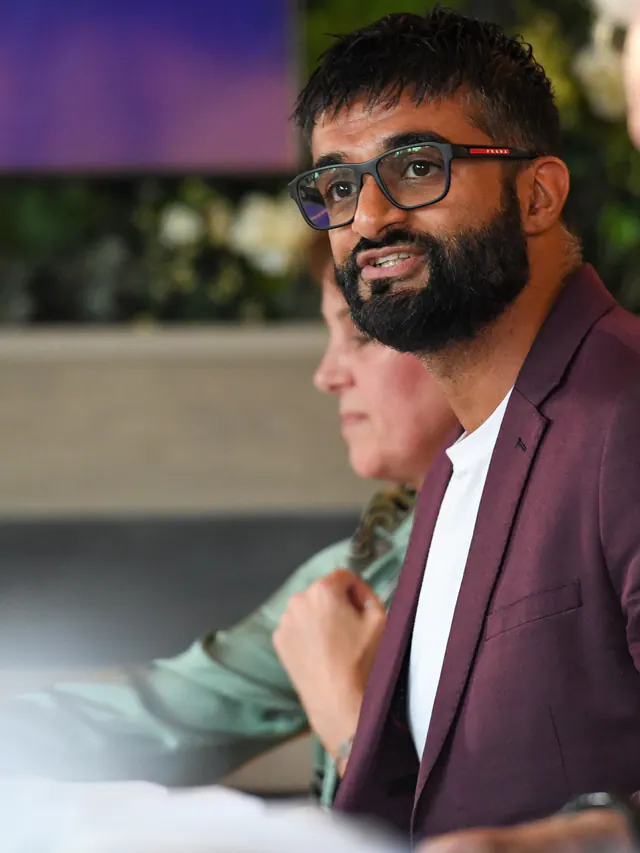 A bearded man in a burgundy blazer and glasses, smiling as he listens across the table.