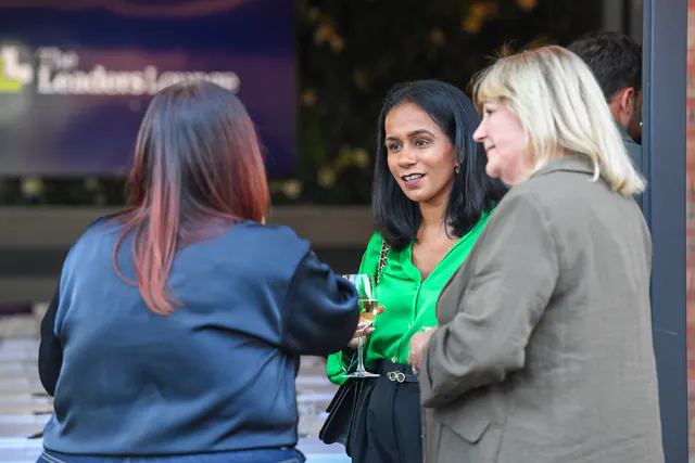 Three women deep in conversation, one holding a glass of wine, with the Leaders' Lounge screen behind them.