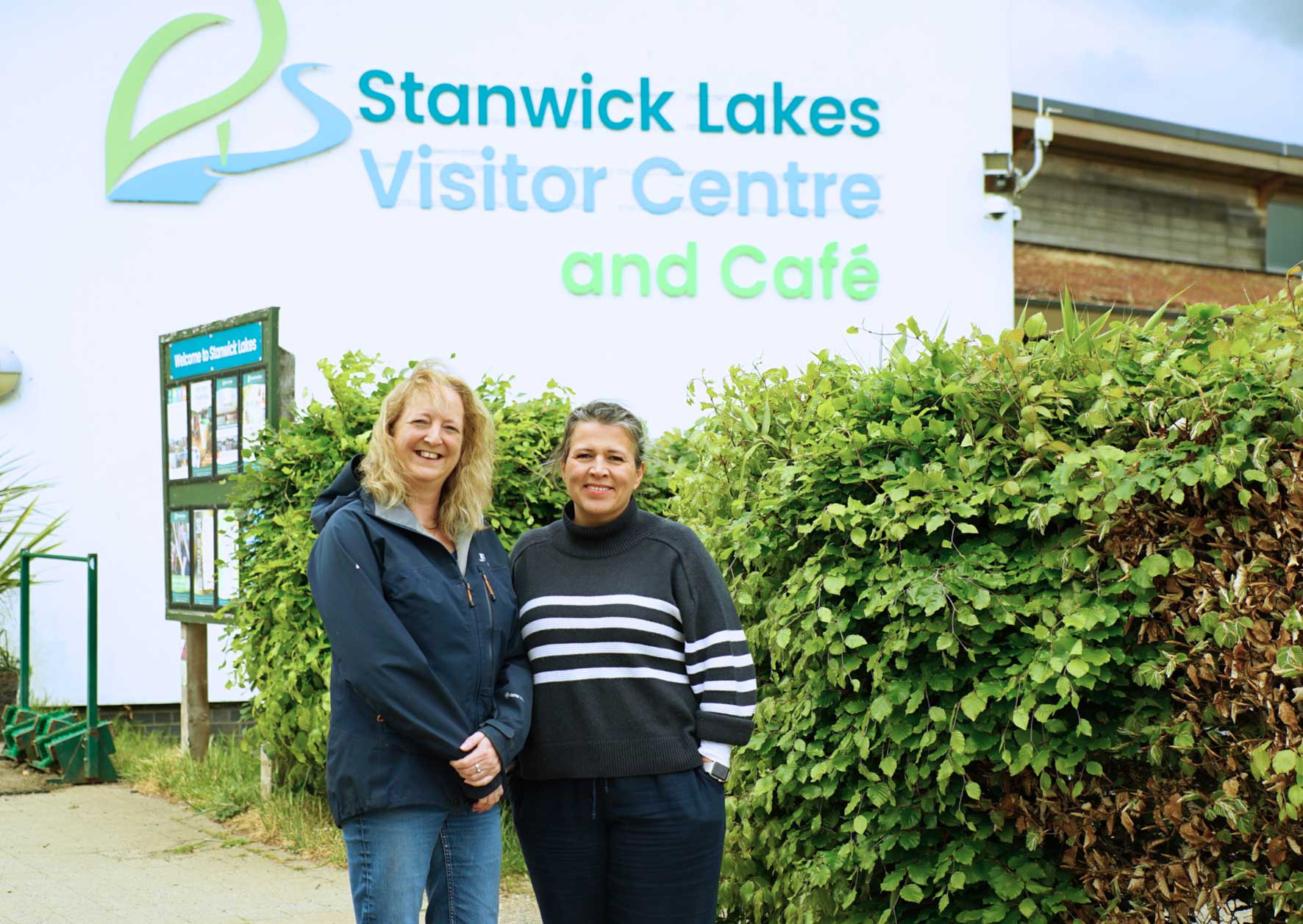 Helen Sillitto (left) and Billie Passchier (right) standing together outside the Stanwick Lakes Visitor Centre and Café, smiling in front of the site's signage and greenery.