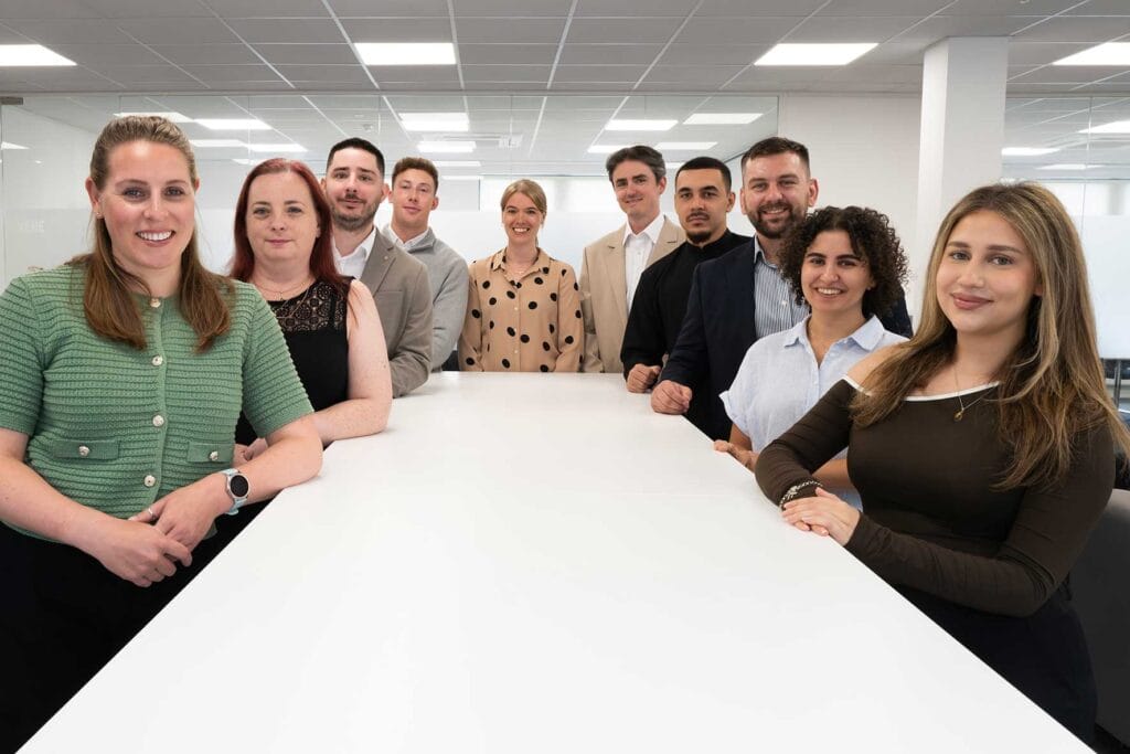 Group of ten professionals posing together around a long white conference table in a modern office, smiling at the camera in business casual attire.