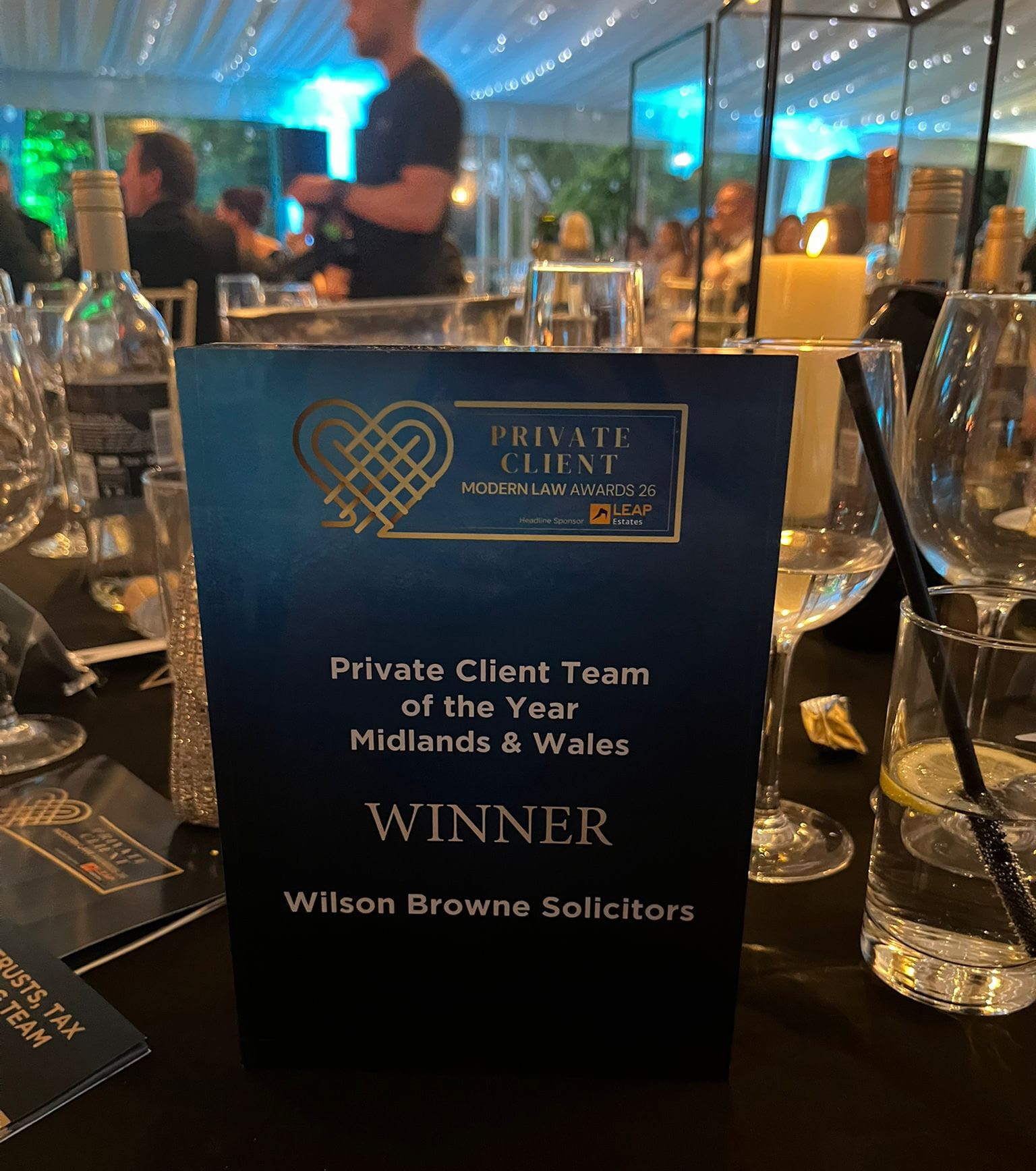 Blue award card on a dining table reading 'Private Client Team of the Year Midlands & Wales' with 'WINNER' and 'Wilson Browne Solicitors'; candle and glassware in a formal event setting.
