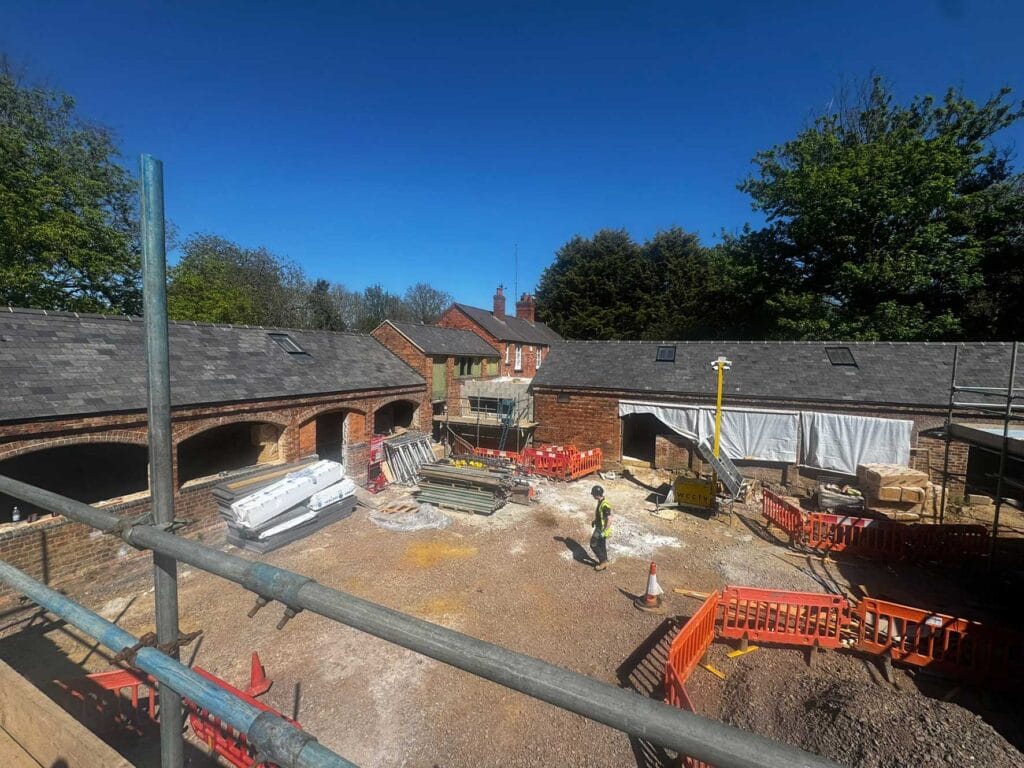 Stables---April-2026 | Business Times Northamptonshire Construction site with brick buildings, scaffolding, and a worker in hi-vis gear amid piles of materials and orange safety barriers on a sunny day.