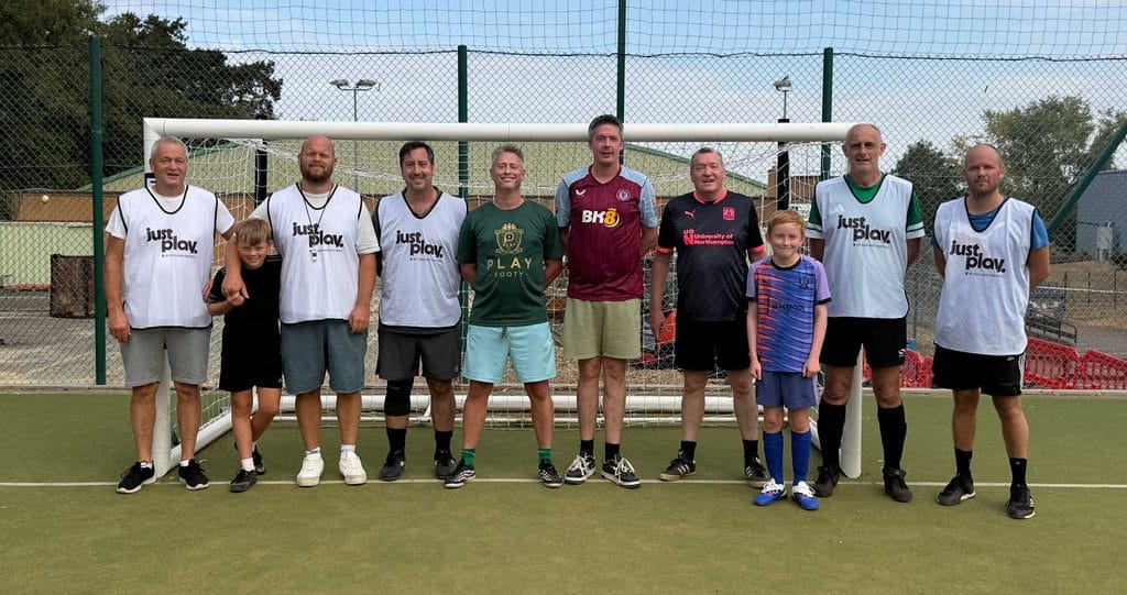 image00003 | Business Times Northamptonshire Group of adults and two kids posing in front of a goal on a football pitch, all wearing casual sportswear and bibs for a team photo.