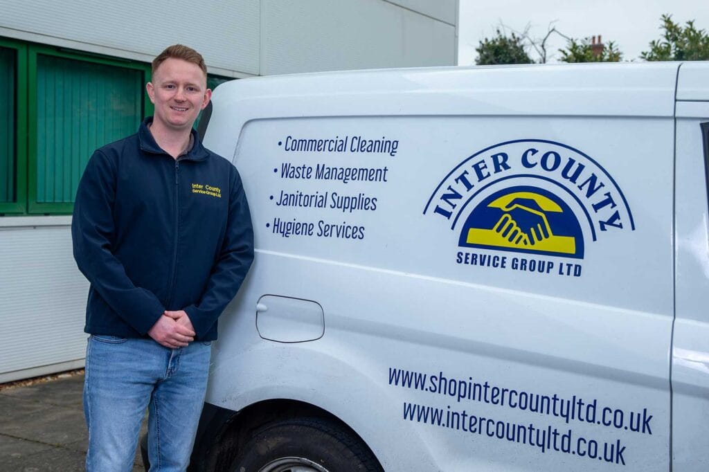 Man in a navy jacket stands beside a white van with the Inter County Service Group logo and a bullet list of services: Commercial Cleaning, Waste Management, Janitorial Supplies, Hygiene Services.