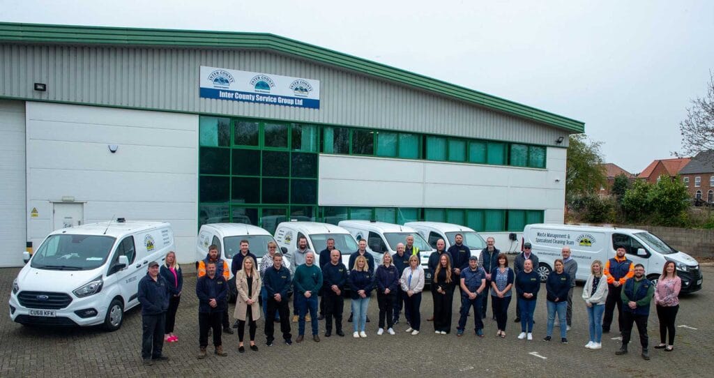 Group photo of employees posing outside a service company building with white vans lined up in front, displaying the Inter County Service Group Ltd sign above.