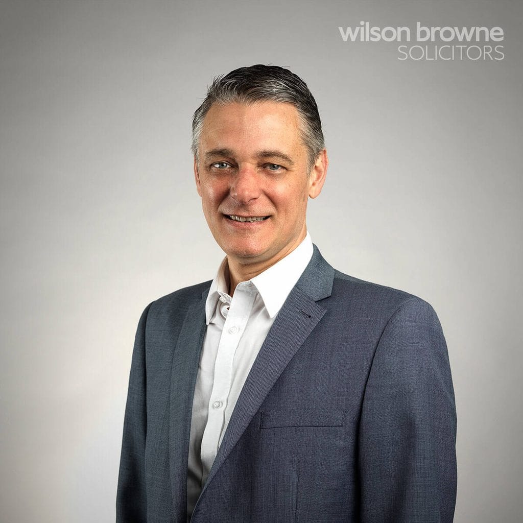 Head-and-shoulders portrait of a smiling man in a gray suit against a neutral background, with the Wilson Browne Solicitors logo in the top-right corner.