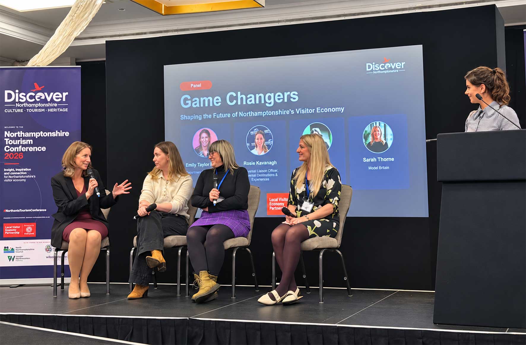 Four women panelists sit on stage and speak into microphones during a panel at the Northamptonshire Tourism Conference 2026, with a large screen behind them showing 'Game Changers'. A Discover Northamptonshire banner stands to the left and a presenter speaks at a podium to the right.