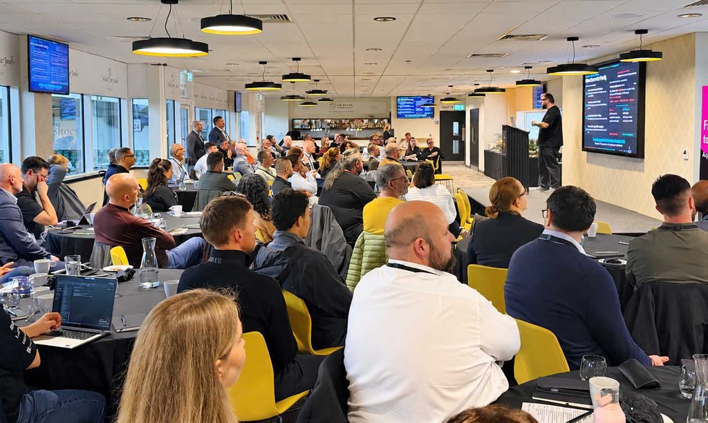 Audience seated at round tables in a bright conference room, listening to a speaker on a stage with large screens displaying information.